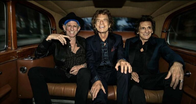 Mick Jagger, Keith Richards e Ronnie Wood seduti sorridenti su un sedile posteriore in pelle marrone di un'auto vintage
