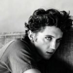 Primo piano in bianco e nero di Troye Sivan con capelli ricci e trucco scuro sugli occhi mentre guarda verso la fotocamera