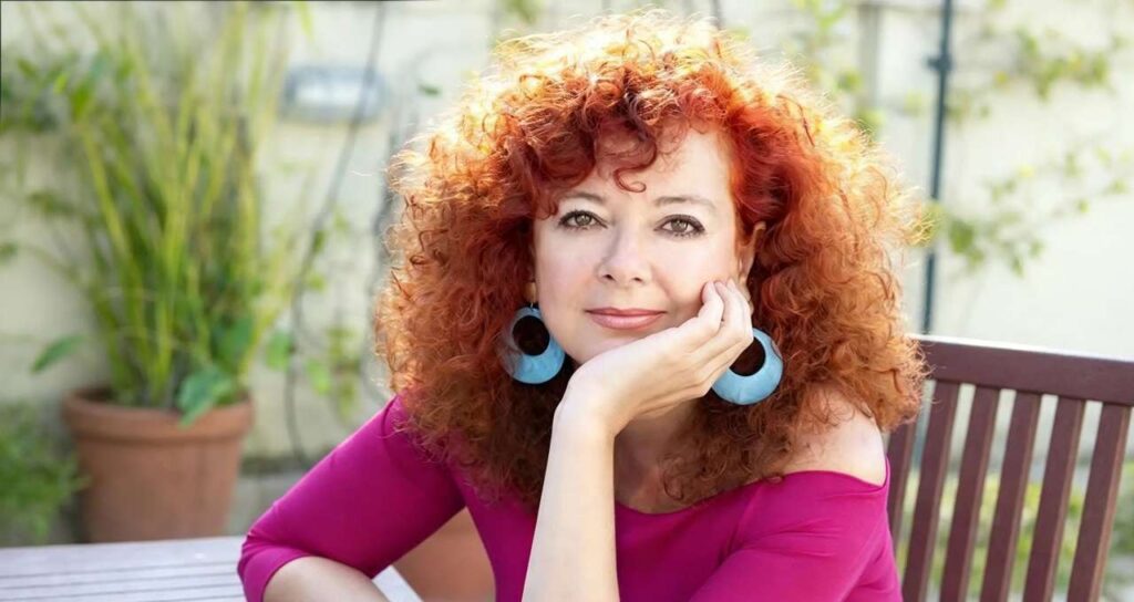 Raffaella Battaglini seduta su una panchina da giardino, capelli ricci rossi, orecchini azzurri e maglia fucsia mentre