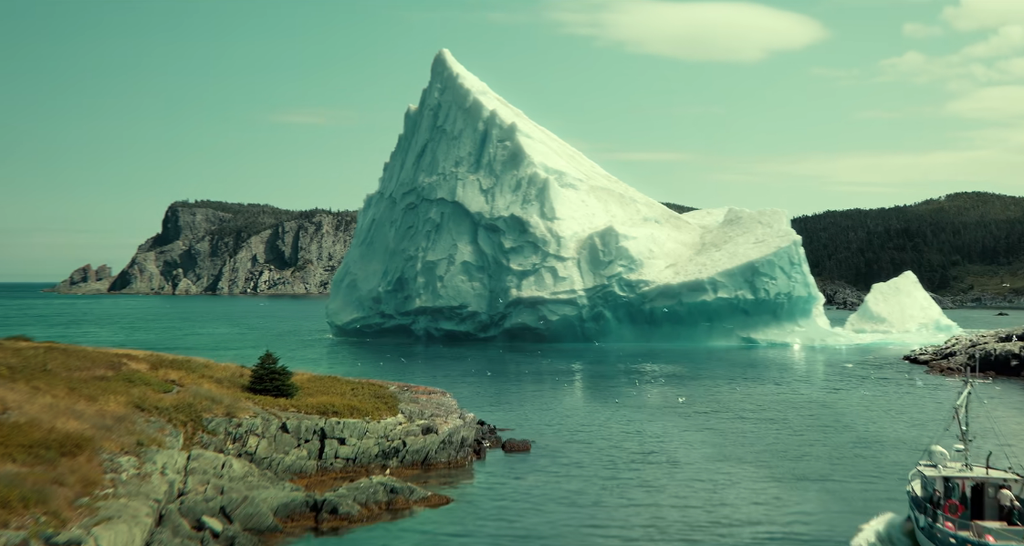 Gigantesco iceberg azzurro vicino alla costa rocciosa con barca a destra e scogliera boschiva sullo sfondo