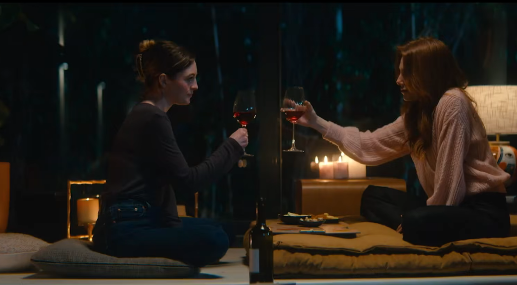 Due donne sedute su cuscini in un soggiorno notturno brindano con calici di vino rosso, bottiglia e candele sul tavolino