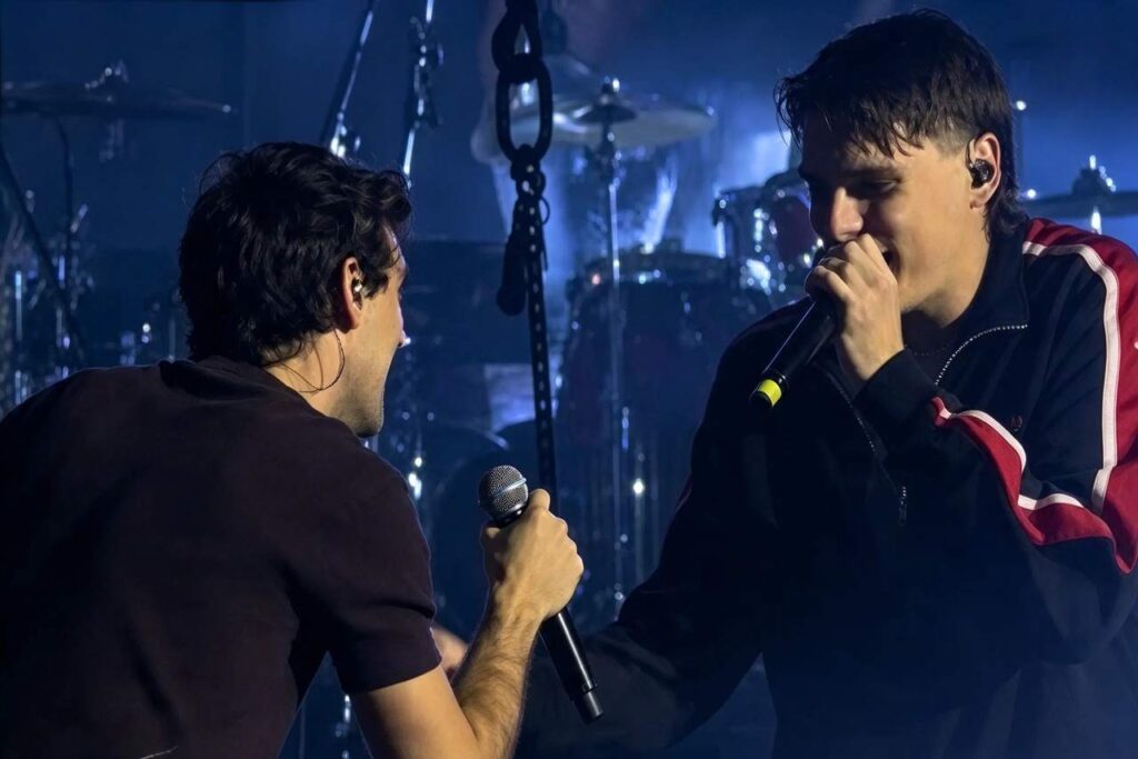Kid Yugi canta al microfono durante un concerto dal vivo, affiancato da un altro artista in un momento di intensa esibizione