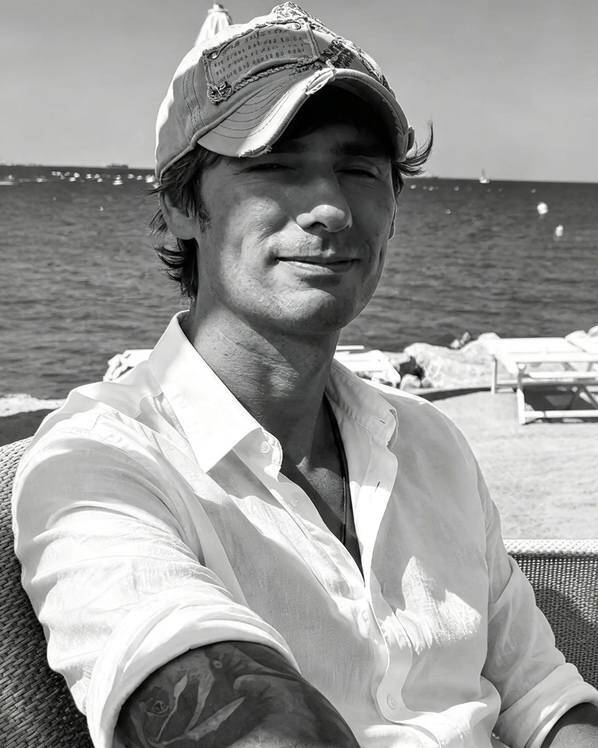 Uomo sorridente con cappellino e camicia bianca seduto su una terrazza vicino al mare, sfondo con sdraio e boe in acqua