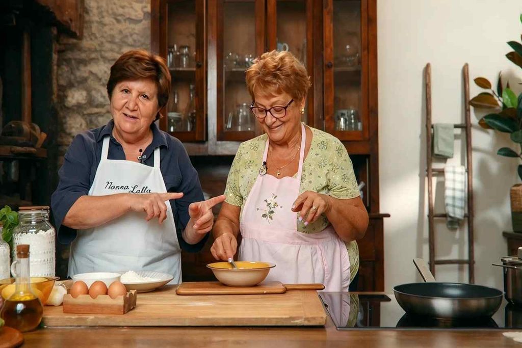 Due signore in grembiule cucinano in una cucina rustica, una indica il grembiule con la scritta Nonna Lalla e l'altra