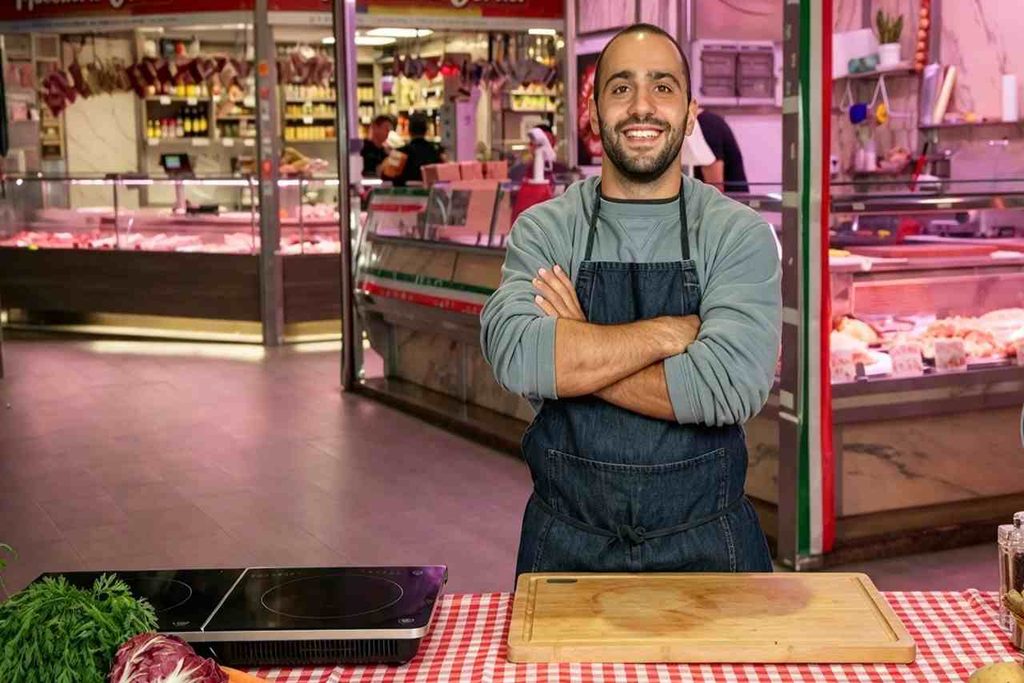 Ruben Bondì sorride con le braccia conserte dietro un bancone con tagliere e piastra a induzione in un mercato di Food