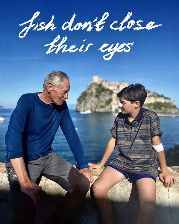 Erri De Luca siede accanto a un ragazzo davanti al mare di Ischia con la scritta Fish don't close their eyes in alto