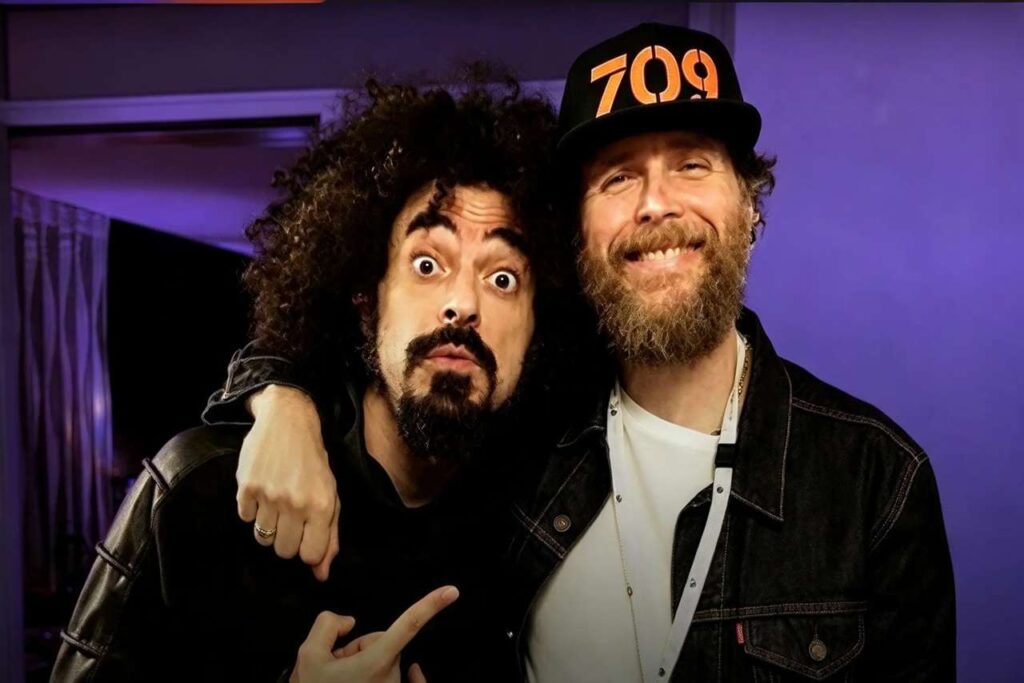 Il cantante Caparezza sorride accanto a un altro uomo che indossa un cappellino con la scritta 709 su sfondo viola
