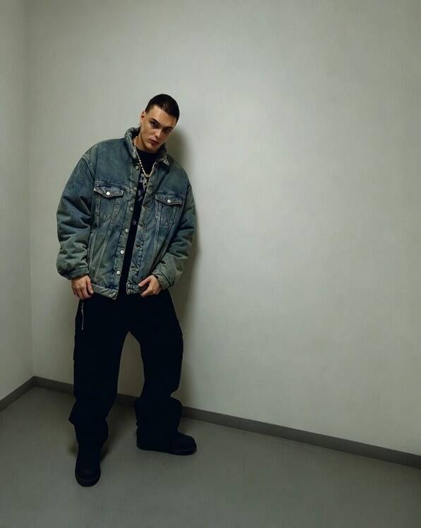 Il rapper Kid Yugi indossa una giacca di jeans oversize e pantaloni neri, posando in piedi contro una parete bianca