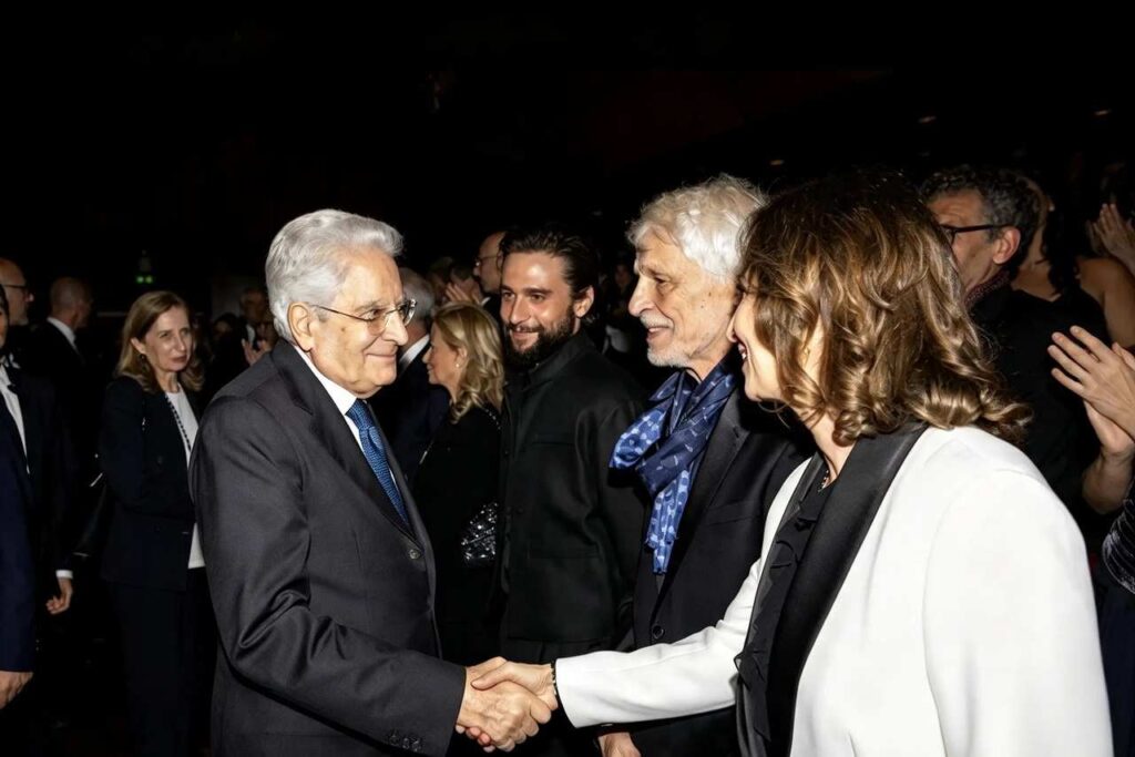 Il Presidente della Repubblica stringe la mano a una donna in giacca bianca in un incontro pubblico, sullo sfondo persone in