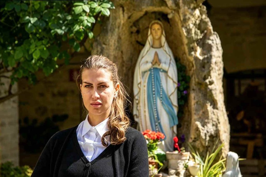Mariana D’Amico in primo piano con camicia bianca e cardigan nero davanti a una statua della Madonna in un giardino del