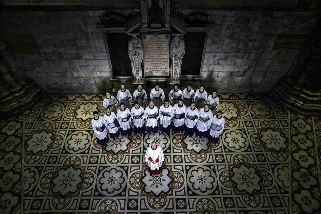 Schola Cantorum Venerandae Fabricae in tonaca bianca e nera disposta a semicerchio su pavimento decorato, direttore Mons