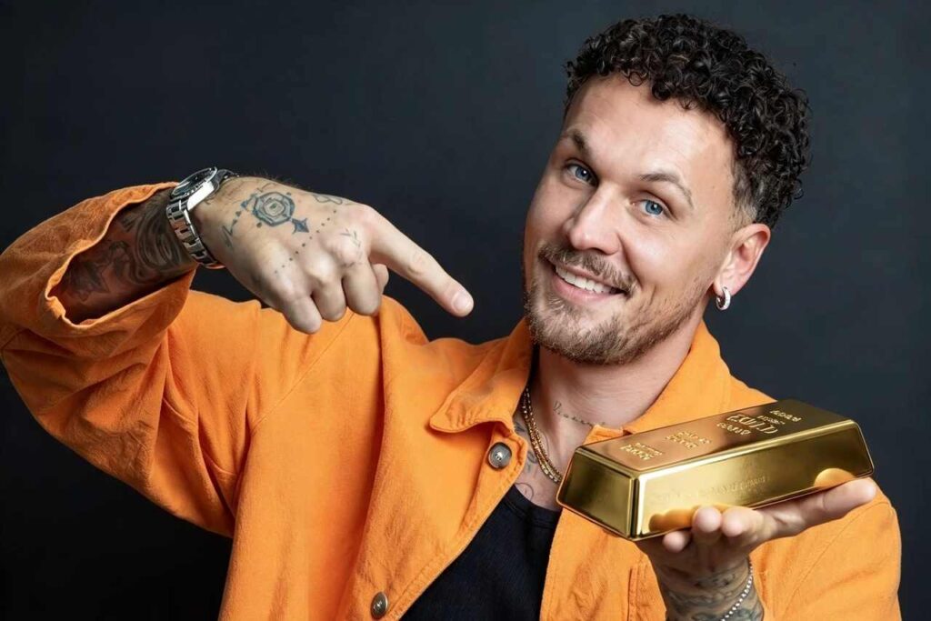 Mr. Oro sorride indossando una giacca arancione mentre indica un lingotto d'oro tenuto nell'altra mano per la nuova stagione