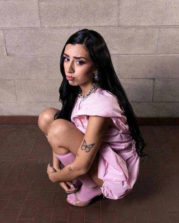 Riri2K accovacciata indossa un vestito rosa e calzini abbinati, mostrando un tatuaggio a forma di farfalla sul braccio