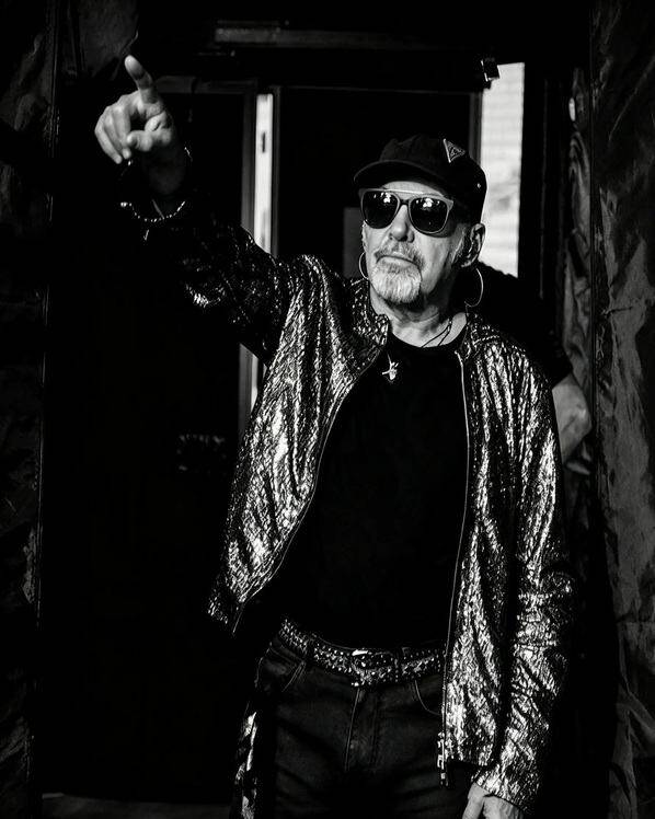 Vasco Rossi in bianco e nero con occhiali da sole e giacca luccicante indica verso l'alto durante il tour Vasco Live 2026