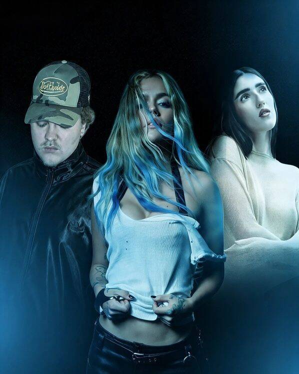 Karol G con capelli blu e top bianco posa tra due persone in un'atmosfera scura e blu per il videoclip di BbY WOW