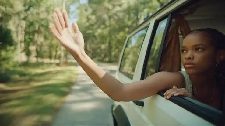 Ragazza afroamericana con braccio fuori dal finestrino di un'auto bianca che viaggia su una strada alberata, sguardo serio