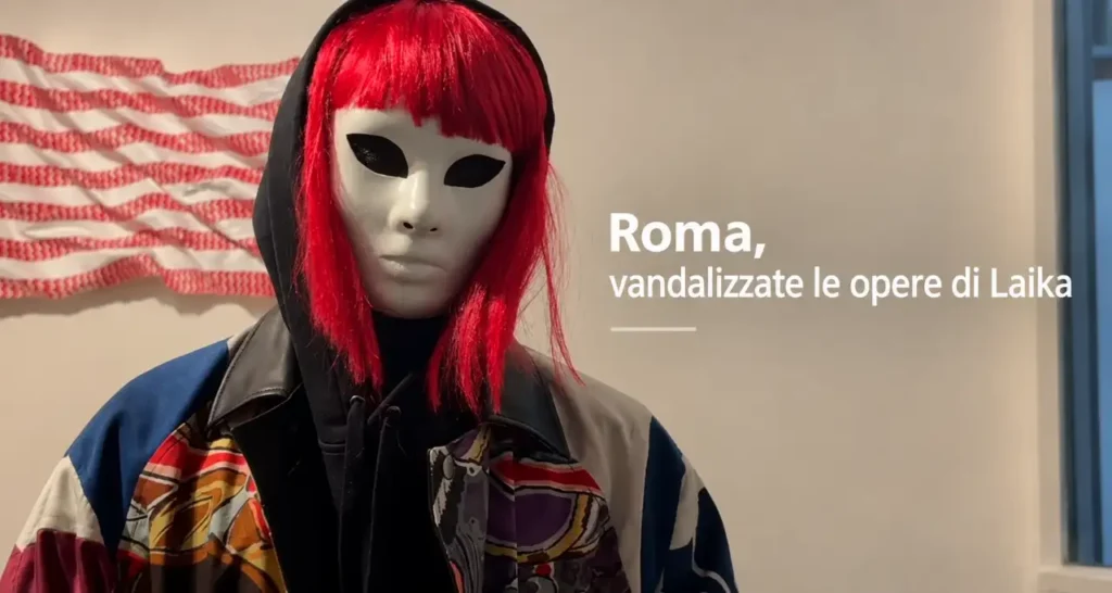 Manichino con maschera bianca, parrucca rossa e giacca multicolore davanti a una parete con la scritta "Roma, vandalizzate
