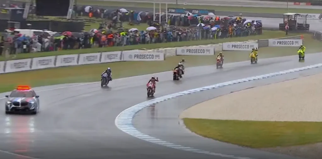 Moto della MotoGP che sfilano su pista bagnata a Phillip Island con auto di sicurezza lampeggiante, piloti in tute colorate