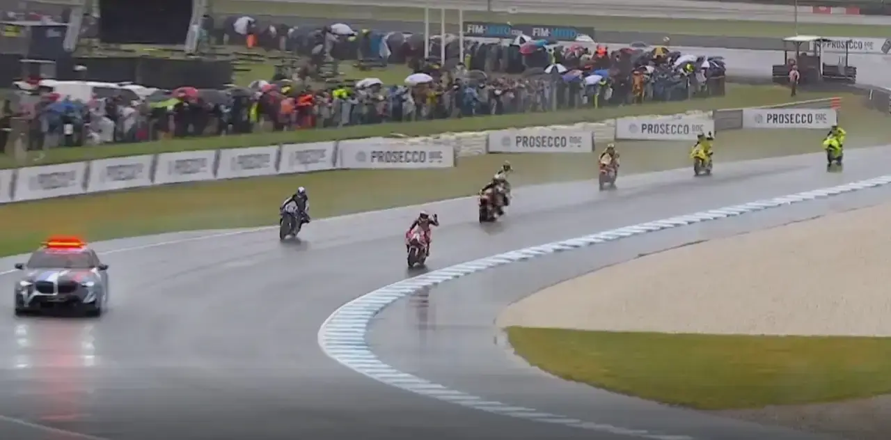 Moto della MotoGP che sfilano su pista bagnata a Phillip Island con auto di sicurezza lampeggiante, piloti in tute colorate
