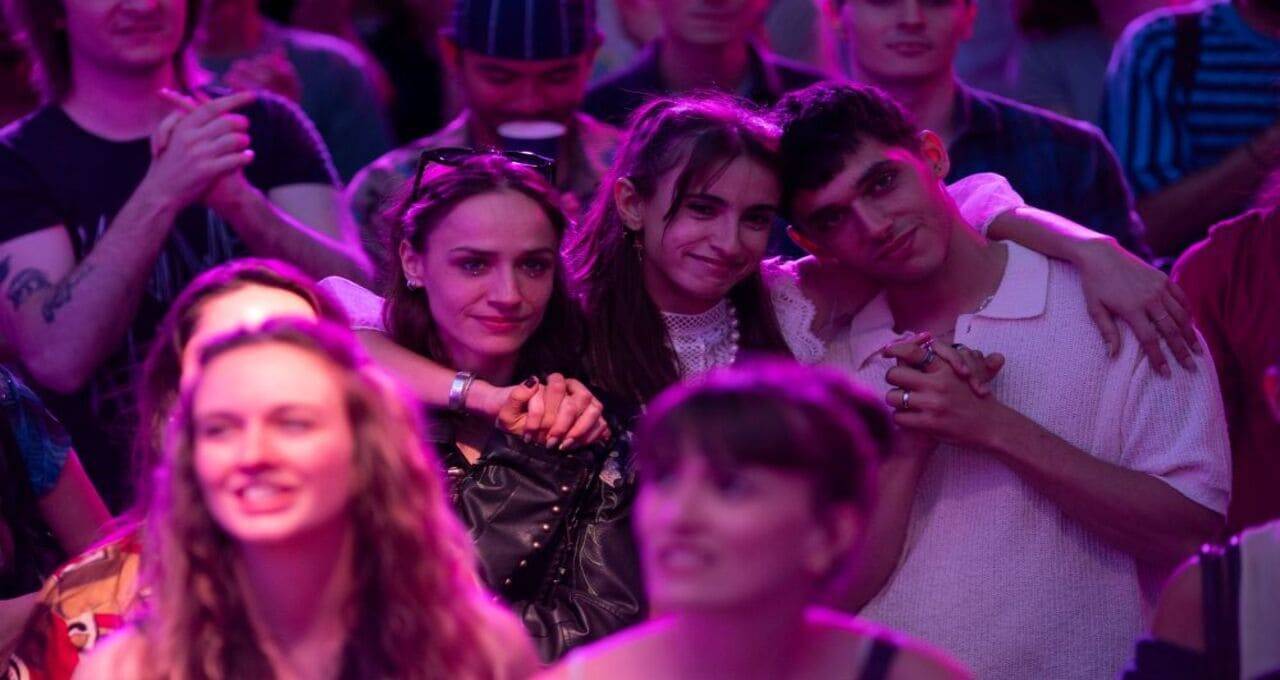 Tre giovani amici sorridenti e abbracciati tra il pubblico in una sala illuminata da luci viola, scena da Il Tempo Di Anne
