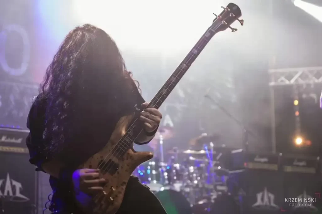 Bassista dai lunghi capelli neri su palco illuminato, tiene un basso marrone e sullo sfondo batteria e amplificatori con il