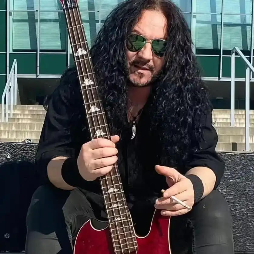 Musicista con capelli lunghi e occhiali verdi seduto sulle scale, tiene un basso rosso e una sigaretta tra le dita, sfondo