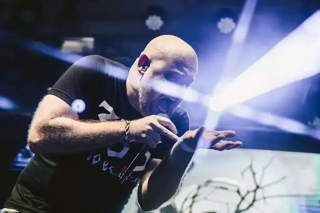 Cantante calvo dei KAT con microfono in mano che urla sul palco illuminato da fari bianchi e blu, maglietta nera con scritta